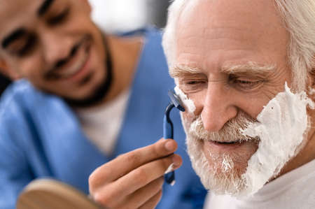 Joyful caregiver shaving an aged man with a razorの写真素材