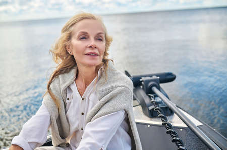 A blonde mature woman in white clothes sitting on a boat and looking dreamyの写真素材