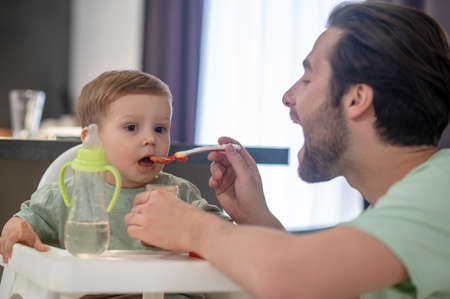 Young father feeding his cute baby son and looking funnyの写真素材