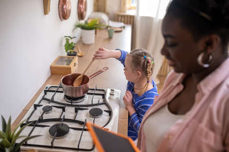 A woman holding a tablet while a girl stirrinf something in a bowl on the gas stoveの写真素材