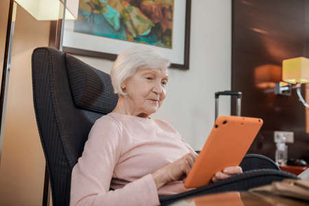 A senior woman in pink blouse with a tablet sitting in an armchair
