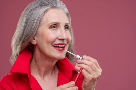 Good-looking mature woman in red putting gloss on her lopsの写真素材