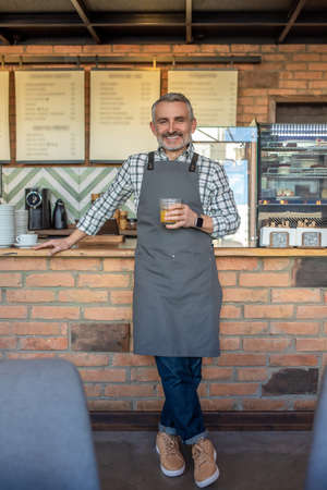 Mid aged man in apron standing near cafe counter with a glass of juice in handの写真素材