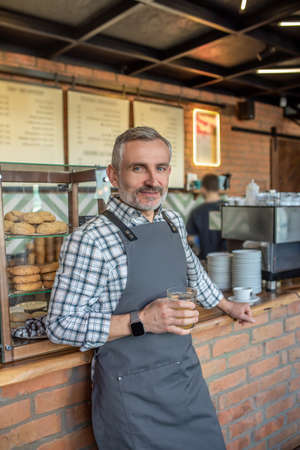 Mid aged man in apron standing near cafe counter with a glass of juice in handの写真素材