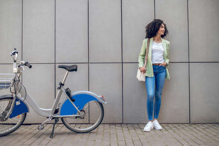 Curly young woman standing near the wall with a bikeの写真素材