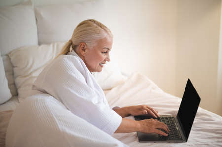 A woman in white robe lying on bed and working on laptopの写真素材