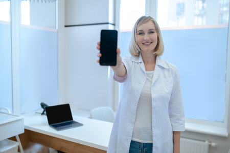 Joyous healthcare worker with the cellphone standing in her officeの写真素材