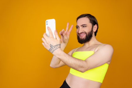 Smiling satisfied bearded transsexual person wearing yellow top posing making selfie showing v signの写真素材