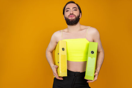 Confident bearded transsexual businessperson wearing yellow top holding folders looking at cameraの写真素材