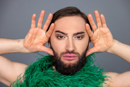 Serious dark haired transsexual person with makeup wearing green boa scarf keeps palms near face
