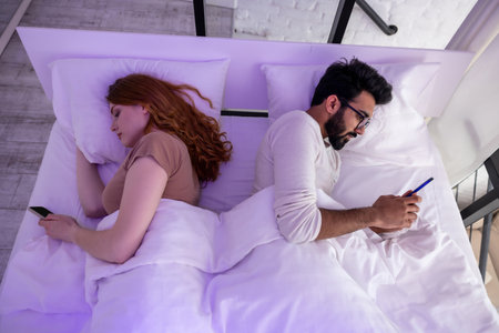 Couple in bed using phone lying backs to each other.の写真素材
