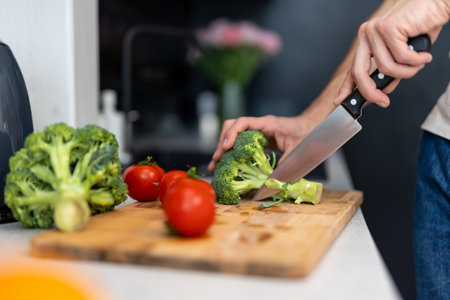 Male hand cuts tomato and brocoli on cutting board with sharp knife in kitchen, making salad