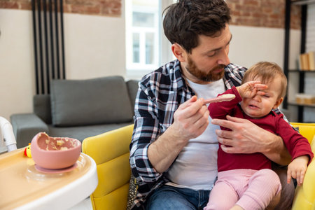 Father feeding crying capricious infant daughter sitting at home.の写真素材