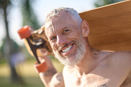 Smiling mature man holding longboard in a city park.の写真素材