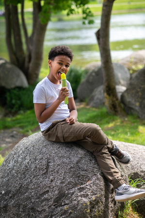 Little boy eating ice cream outdoors on hot summer day, sitting on rock, enjoying sweet dessert.の写真素材