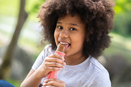 Little girl with curly hair eating delicious chocolate ice cream in garden and enjoys on hot day.の写真素材