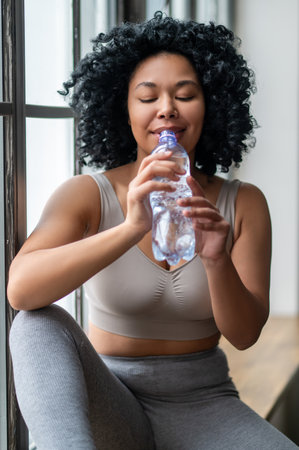 Contented curly-haired woman in sportswear sitting on the wondow with bottle of water