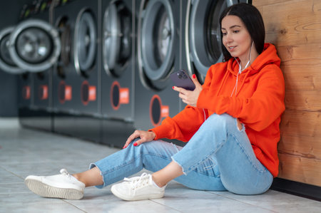 Young woman listening music with smartphone and earphones waiting in laundry room.の写真素材