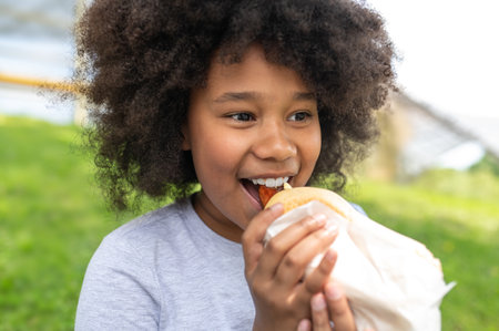 Dark skinned female child eating junk food with pleasure in park while sitting on green grass.の写真素材