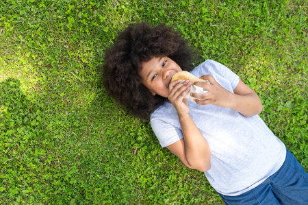 Girl lying on grass and eating fast food in park, happy kid having snack after using laptop outdoor.