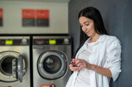 Woman waiting in a laundry room using mobile phone, browsing internet.の写真素材