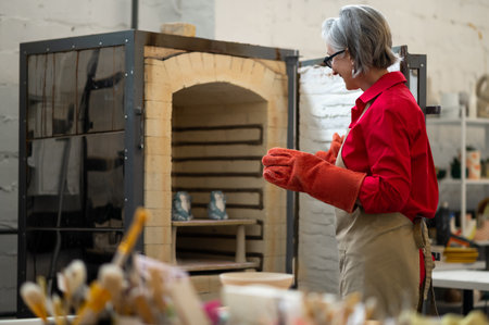 Mature craftswoman taking clay products from pottery kiln.