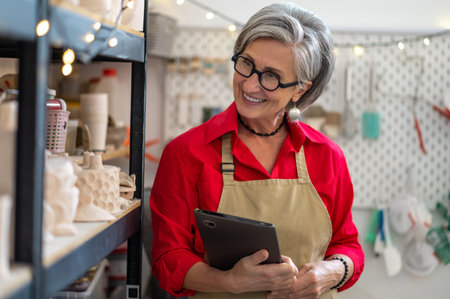 Happy female ceramist using digital tablet in store, managing store with handmade ceramic productsの写真素材