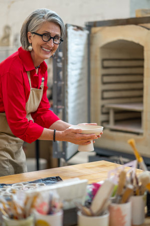 Craftswoman holding clay plate before placing into kiln for firing pottery while working in workshopの写真素材