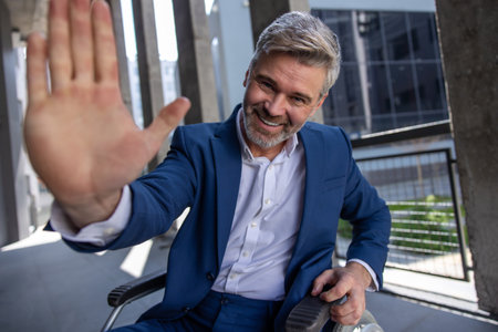 Gray haired disabled man wearing official style suit in wheelchair, raising palm, giving high fiveの写真素材