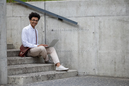 Curly-haired young man in white shirt sitting on the steps with laptop