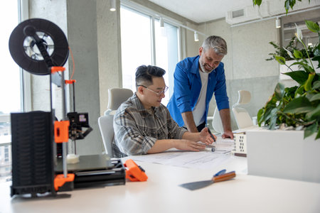 Men working with 3D printer and blueprint for designing mechanical parts in high tech workshop.