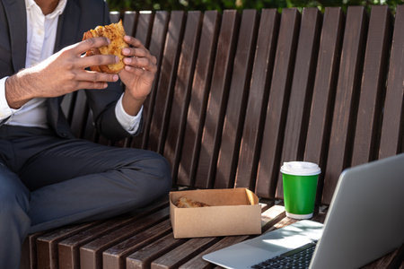 Young man having lunch and working on laptop while sitting on the bench outsideの写真素材
