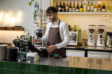 Male barista at counter using cashbox computer in cafe store checking clients order