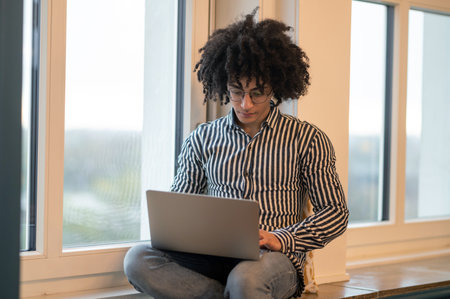 Curly-haired man sitting on a window-sill and working on laptop