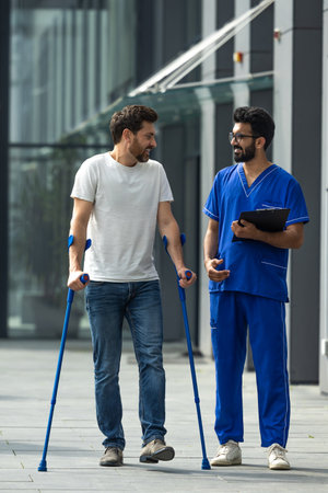 Young bearded man with crutches walking and talking to a male nurse