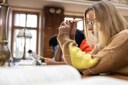 Blonde young woman sitting at the table in the library and feeling tiredの写真素材