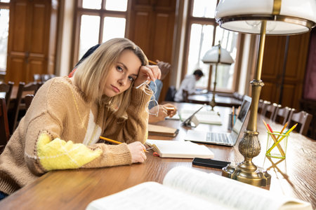 Blonde young woman sitting at the table in the library and feeling tired
