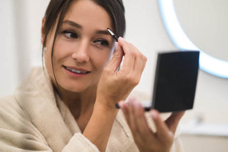 Pretty young woman looking in the mirror and making her eyebrows