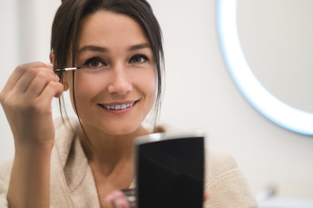 Cute dark-haired young woman doing her make up and looking contented