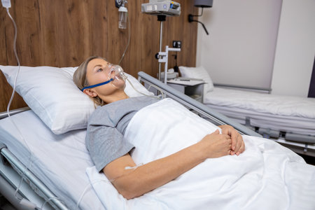 Blonde mid aged woman lying in bed in hospital with an oxygen mask on her faceの写真素材