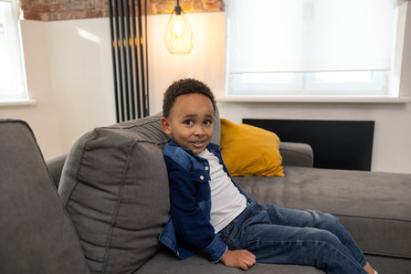 Cute charming black boy sitting on comfortable sofaの写真素材