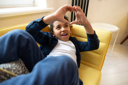 Cute boy showing heart shape with hands while sitting on a sofa at homeの写真素材