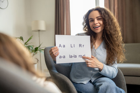 Cute smiling young speech therapist with table with written syllables in handsの写真素材