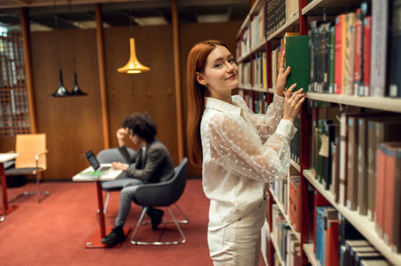 Cute young ginger girl in white with a book in hand standing near the bookshelfの写真素材