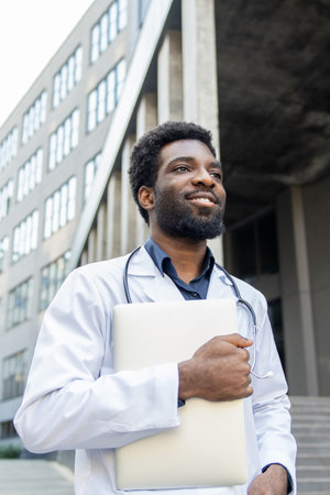 Smiling doctor holding laptop in hands standing outdoor modern hospitalの写真素材