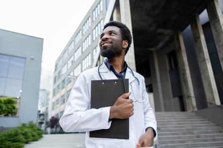 Joyful doctor in medical gown holding clipboard filled with notes standing outdoors hospitalの写真素材