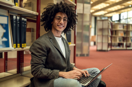 Curly-haired young man sitting on the floor in the library and working on laptop