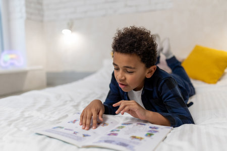 Curly-haired african american kid lying on the bed with a book and looking excitedの写真素材