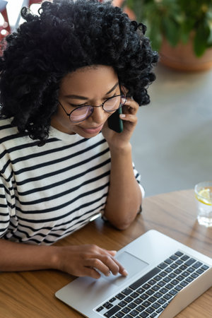 Curly-haired woman working on laptop and having a phone callの写真素材