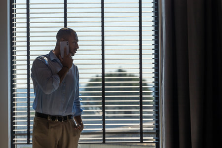 Man wearing shirt posing near big window in office and talking on mobile phoneの写真素材